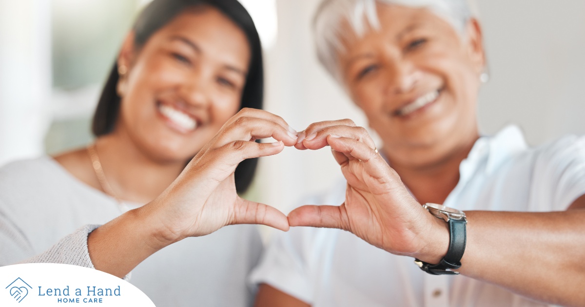 A daughter and an older mother create a heart with their hands, representing the feeling that is highlighted during National Family Caregivers Month